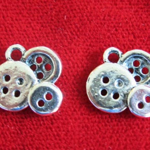 May include: Two silver charm pendants shaped like buttons. Each pendant features three buttons, one large and two smaller buttons.