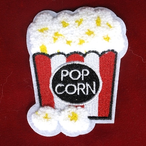 May include: A fabric patch shaped like a popcorn bucket with red and white vertical stripes. The top features textured white and yellow popcorn, and a central black circular emblem displays the white text POP CORN in a bold, sans-serif font.