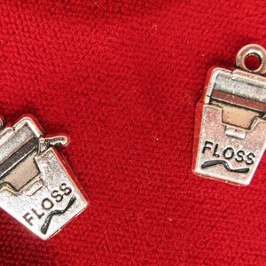 May include: Two silver-colored charms shaped like floss containers, each with the word "FLOSS" in black lettering. The charms have a small loop at the top for attaching to a necklace or bracelet. The background is a textured red fabric.