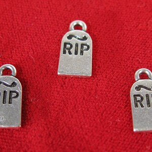 May include: Three silver-toned metal charms shaped like tombstones with the word "RIP" engraved on them.