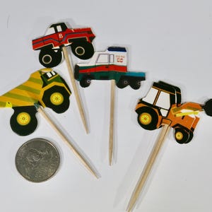 May include: Four colorful construction vehicle cupcake toppers. The toppers feature a red truck, a yellow dump truck, a blue and white delivery truck, and an orange tractor. Each topper is attached to a wooden stick.