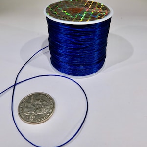 May include: A spool of dark blue elastic cord with a silver coin for scale. The cord is wound around a white plastic spool with a holographic label.