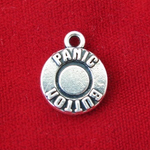 May include: A silver-colored charm with the words "PANIC BUTTON" engraved around a central circle. The charm has a loop at the top for attaching to a necklace or bracelet. The background is a solid red fabric.