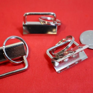 May include: Three silver-colored metal hardware clips for keychains, each featuring a rectangular clamp with an attached split ring. A standard United States quarter coin is placed next to the hardware to provide a visual scale reference.