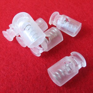 May include: A collection of clear plastic cord locks, each featuring a cylindrical body with a spring-loaded mechanism inside. These small fasteners are designed to secure drawstrings on clothing or gear, showing a translucent finish.