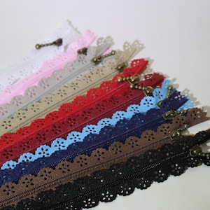 May include: A collection of decorative zippers in various colors, including white, pink, gray, red, blue, navy, brown, and black. Each zipper features a scalloped edge and a bronze-colored pull.