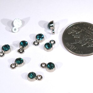 May include: A close-up of eight teal blue rhinestone charms with silver loops. The charms are arranged on a white surface next to a US quarter.