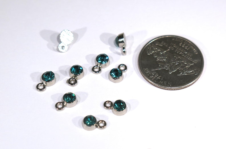 May include: A close-up of eight teal blue rhinestone charms with silver loops. The charms are arranged on a white surface next to a US quarter.