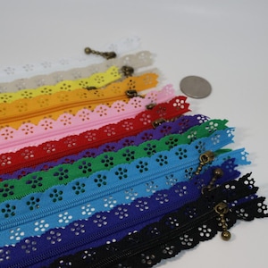May include: A collection of multi-colored zippers featuring a decorative scalloped edge with a floral eyelet pattern. Each zipper includes a bronze-toned metal pull tab. A small silver-colored coin is placed near the zippers for scale reference.