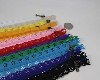 May include: A collection of multi-colored zippers featuring a decorative scalloped edge with a floral eyelet pattern. Each zipper includes a bronze-toned metal pull tab. A small silver-colored coin is placed near the zippers for scale reference.