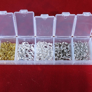 May include: A clear plastic storage box with eight compartments filled with gold, silver, and brass jump rings. The box is on a red surface.
