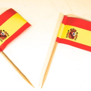 May include: Two small Spanish flags with red and yellow stripes and a coat of arms. The flags are on wooden sticks, set against a white background. The flags are likely used for decoration or to represent Spain.