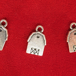 May include: Three metal barn charms with a rustic finish. Each charm features a barn shape with a door design and a loop for attaching to jewelry or crafts. The charms are displayed on a red fabric background.