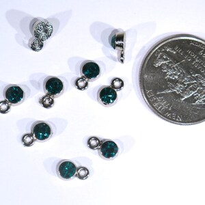 May include: A close-up of ten silver-toned metal charms with dark green rhinestones. The charms have a loop on one end and a small, round rhinestone on the other end.