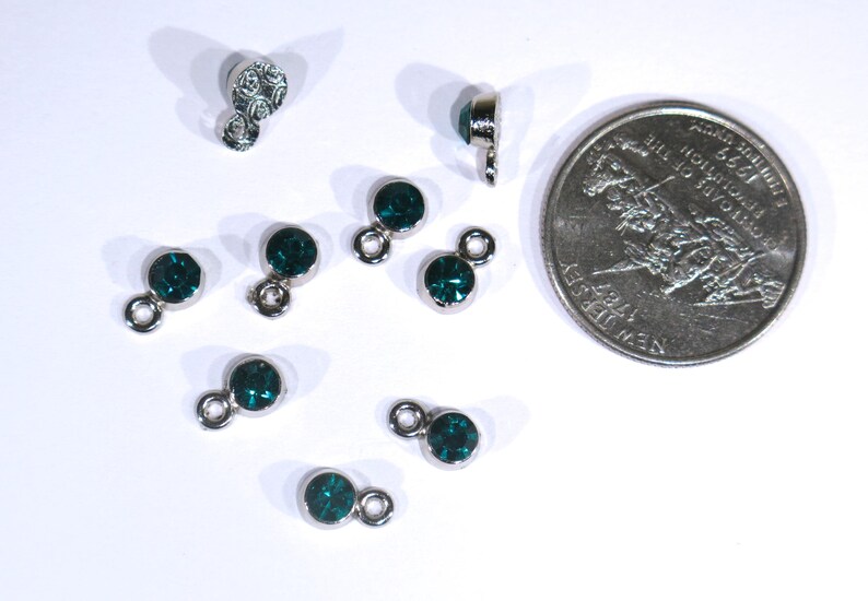 May include: A close-up of ten silver-toned metal charms with dark green rhinestones. The charms have a loop on one end and a small, round rhinestone on the other end.