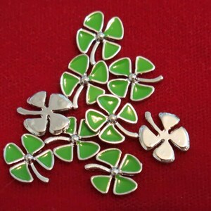 May include: A group of nine silver four-leaf clover charms with green enamel. The charms are scattered on a red surface.