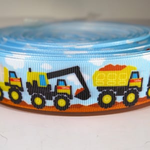 May include: A roll of blue ribbon featuring a repeating pattern of construction vehicles. The ribbon has a light blue background with white clouds. The construction vehicles are yellow, black, and orange.
