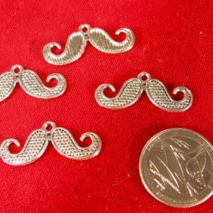 May include: Silver-colored mustache charms with textured details, displayed on a red background. A coin is included for size reference. These charms are suitable for jewelry making or craft projects.
