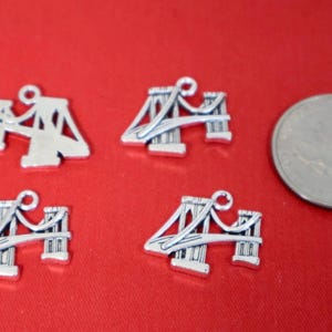 May include: Four silver-toned metal charms shaped like suspension bridges, each featuring a small loop at the top for attachment. A standard United States quarter coin is positioned to the right for scale, measuring approximately 0.955 inches in diameter.