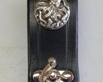 May include: Black leather belt loop with a silver-colored octopus button and a silver clasp. The belt loop is designed to hold a knife or other small item. The button is round and has a detailed octopus design.
