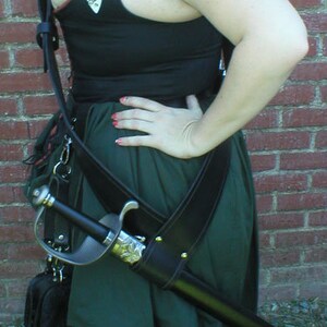 May include: A woman wearing a black vest with a green skirt and a leather sword scabbard. The vest has several decorative patches and the sword is strapped to the belt.