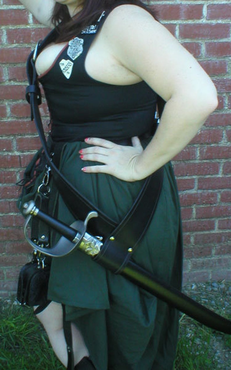 May include: A woman wearing a black vest with a green skirt and a leather sword scabbard. The vest has several decorative patches and the sword is strapped to the belt.