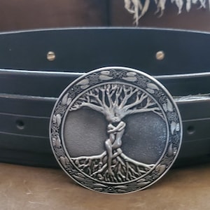May include: A black leather belt with a silver buckle featuring a tree of life design. The buckle is ornate and detailed, with a pair of figures intertwined within the branches of the tree.
