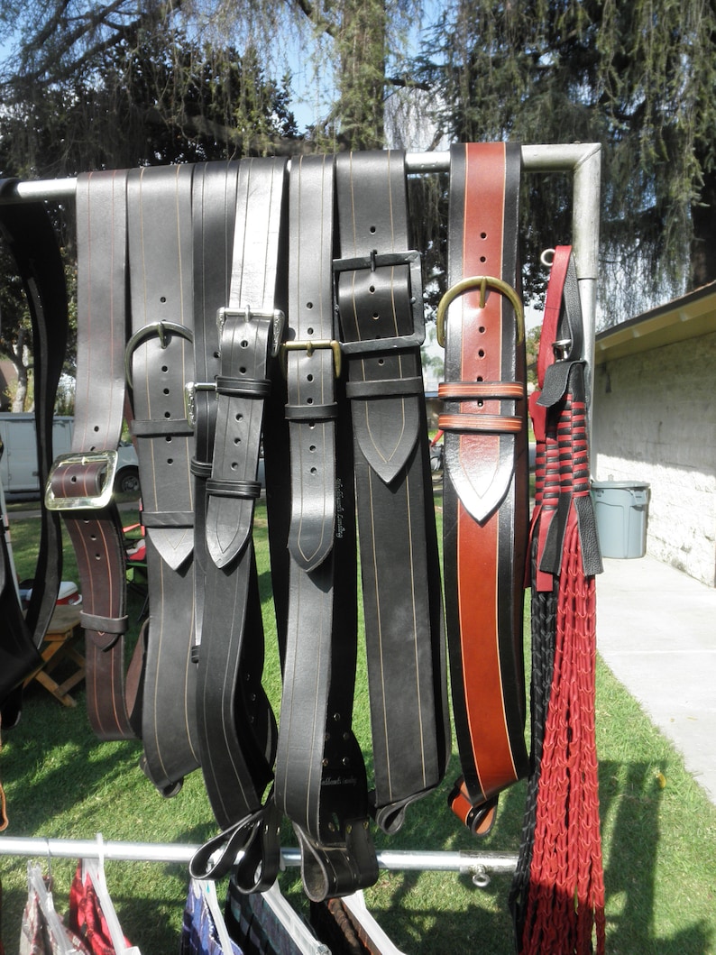 May include: A collection of eight leather belts hanging on a metal rack. The belts are various shades of brown and black, with some having decorative stitching.