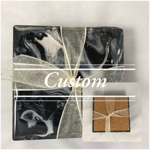 May include: Set of four black and white marble coasters with a silver glitter ribbon tied around them. The coasters are made of resin and have a smooth, glossy finish. The word "Custom" is written on the ribbon.
