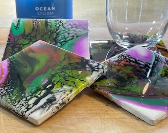 May include: Four square coasters featuring abstract artwork in shades of green, black, pink, and white. The coasters are made from a smooth, glossy material.