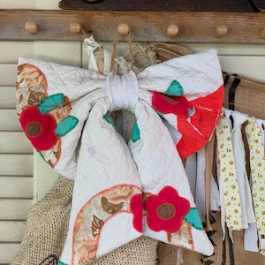 May include: A large decorative bow crafted from white quilted fabric featuring appliqued floral motifs in bright red, brown, and teal. The bow has long, flowing tails and is constructed with visible stitching, showcasing a textured surface and a central knot.