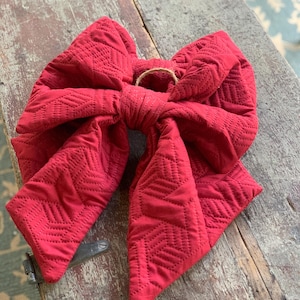 May include: A large, deep red fabric bow featuring a quilted, geometric herringbone pattern throughout. The bow is tied in a central knot and includes a small, circular jute twine loop attached at the top center for hanging purposes.