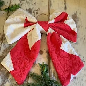 May include: A decorative bow made from quilted fabric featuring a red and white geometric pattern. The bow has two large loops and two long, pointed tails, with a small twine loop attached at the center for hanging purposes.