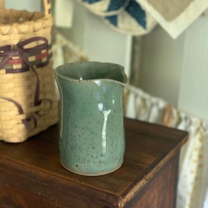 May include: A sage green ceramic pitcher with a speckled glaze finish. The vessel features a cylindrical body with a slightly flared rim and a small integrated pouring spout. The surface has a smooth, glossy texture with visible dark flecks.