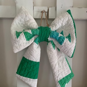 May include: A large decorative bow constructed from quilted fabric featuring a white base with green geometric patterns and solid green accents. The bow is tied in the center with a green fabric knot and includes a thin twine loop for hanging purposes.