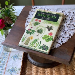 Op de afbeelding: Een hardcover boek getiteld The Woman's Day Book of HOUSE PLANTS door Jean Hersey, met een crèmekleurige kaft met kleurrijke botanische illustraties van diverse potplanten, waaronder een cactus, geraniums en groen blad.