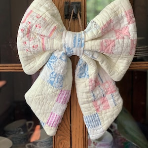May include: A large decorative bow crafted from quilted fabric featuring a patchwork design. The quilt pattern consists of various squares in soft shades of cream, pale pink, and light blue, with floral and striped prints. The bow is tied with a center knot.