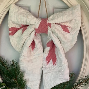 May include: A decorative bow made of quilted white fabric with a pink patterned design. The bow features a central knot and two long, hanging tails, and it is suspended by a thin brown twine loop for hanging purposes.