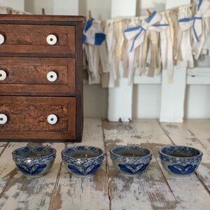 May include: Four small, round ceramic bowls with a blue floral design. The bowls are arranged on a distressed white wooden surface. A dark wooden cabinet with white knobs is in the background.