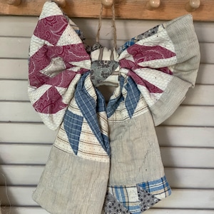 May include: A large decorative bow crafted from quilted fabric patches. The design features a mix of patterns including burgundy paisley, blue plaid, and neutral pinstripes. The bow is constructed with various textile segments stitched together.