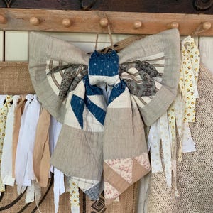 May include: A decorative fabric bow constructed from various quilted textile scraps in shades of blue, beige, cream, and brown. The bow features a mix of striped, floral, and solid patterns with a central knot and two long, hanging fabric tails.