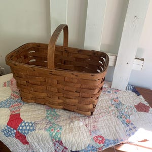 May include: A rectangular, woven brown basket with a wooden handle. The basket is sitting on a vintage quilt with a patchwork design of red, white, and blue circles. The basket is empty.