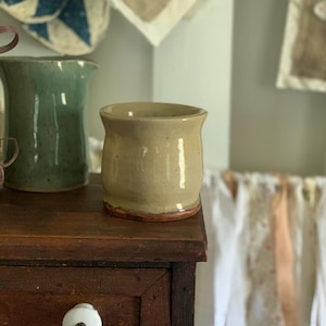 May include: A small, cylindrical ceramic cup with a smooth, glossy beige glaze and a visible reddish-brown clay base. The cup features a slightly flared rim and a rounded body, sitting alongside a taller, speckled sage green ceramic pitcher.