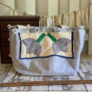 Op de afbeelding: Een lichtgrijze canvas draagtas met een rechthoekige gewatteerde patch met een crème, groen en grijs bloemmotief. De tas heeft metalen ringen langs de bovenrand en staat achter een houten liniaal van 60 cm lang.