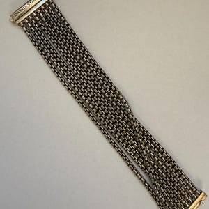 May include: Silver-toned bracelet with multiple strands of small, rectangular links. The bracelet has a rectangular clasp with the engraved text "DAVID YURMAN". The jewelry is displayed on a light-colored surface.