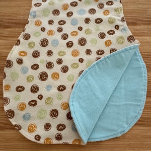 Op de afbeelding: Een baby slabbetje met een crèmekleurige voorkant met bruine, gouden, groene en blauwe spiraalvormige patronen. De achterkant van het slabbetje is lichtblauw. Het slabbetje is gemaakt van zachte stof en heeft een gebogen onderkant.