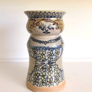 May include: A cylindrical ceramic vase with a speckled beige glaze featuring a hand-painted illustration of a figure with curly hair, a necklace, and a floral-patterned garment. The design uses dark blue and yellow outlines and accents.