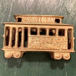 May include: Gold-colored metal miniature replica of a San Francisco cable car. The cable car has the words "Cable Car San Francisco" on the side.