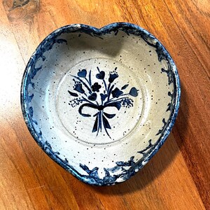 May include: Heart-shaped ceramic bowl with a blue floral design. The bowl has a speckled, off-white interior and a blue, scalloped rim. A blue floral bouquet is centered inside the bowl. The bowl is sitting on a wooden surface.