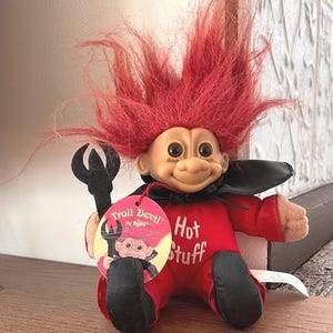 May include: A Troll doll with red hair, wearing a red outfit with the words "Hot Stuff" and a black cape. The doll holds a black trident and has a tag that says "Troll Devil by RJSS".